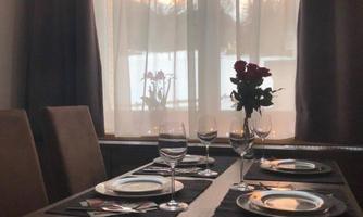 An elegantly set table with dishes and glasses, decorated with a vase of red roses. In the background, there is a beautiful view of nature through a large window.