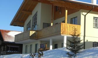 A modern house with a wooden balcony, surrounded by snow. The architecture is clear and friendly, typical of alpine regions.