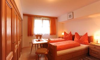 A cozy bedroom with two single beds and orange pillows. The room features wooden wardrobes and a small table, complemented by warm window decorations.
