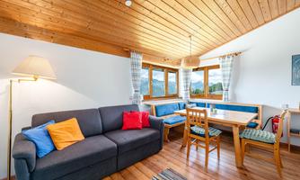 A cozy living space with a gray couch and colorful cushions. Large windows offer a view of the surrounding nature.
