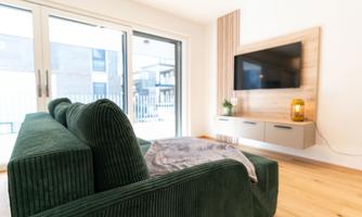A modern living room with a green sofa and a TV on the wall. Large windows let in plenty of daylight and offer a view outside.