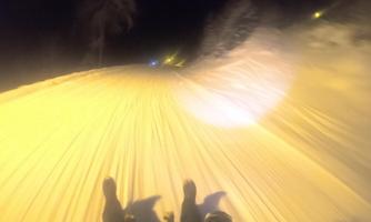 A snow-covered slope at night, illuminated by yellow lights. The view shows a dynamic descent with snow in focus.