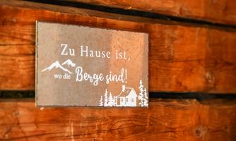 A sign with the inscription "Home is where the mountains are!" hangs on a wooden wall. It features an alpine landscape with mountains and a small house.