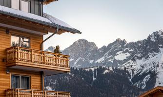 A wooden house with a balcony and a person enjoying the view. In the background, snow-covered mountains can be seen.