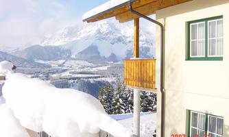 A snow-covered village with a wooden balcony and modern buildings. Majestic mountains rise in the background under a clear sky.