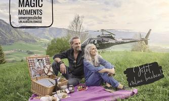 A couple is sitting on a blanket in the greenery and enjoying a picnic. In the background, there is a helicopter, while the landscape offers a beautiful view.