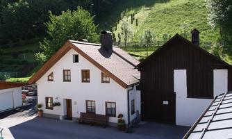 A traditional house with a white and a dark wood part. It is located in a green environment with trees and hills.