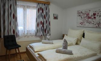 A cozy bedroom with two beds and a chair. The windows are decorated with curtains, and a floral painting hangs on the wall.