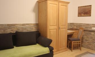A cozy room with a wooden wardrobe and a couch with a green blanket. A table with a chair is also present in the room.