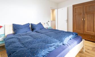 A modern bedroom with a large bed and blue bedding. In the background is a wooden wardrobe and a bright wall.