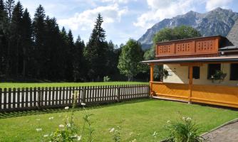 A cozy house with a well-kept garden and a fence. In the background, tall trees and impressive mountains can be seen.