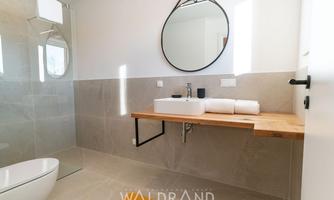 A modern bathroom with bright marble and a large round mirror. The wooden shelf provides space for towels and cosmetics.