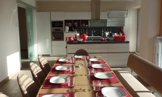 A modern dining area with a long wooden table set for a meal. The kitchen in the background is bright and well-equipped.