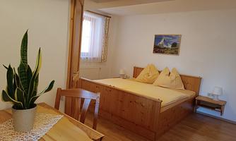 A cozy bedroom with a double bed and bright pillows. A table with a plant and wooden floor create an inviting atmosphere.