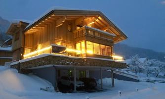 A warmly lit wooden house in a snow-covered landscape. The sky is gray and snowflakes are falling.