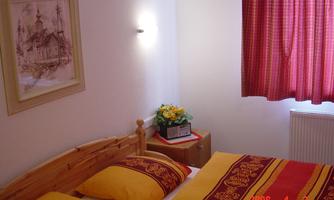 A cozy bedroom with a double bed and yellow cushions. The room has a red curtain and a small flower decoration on the nightstand.