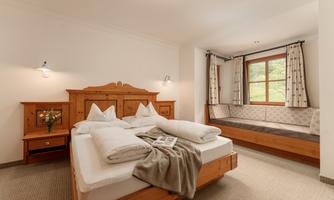 A cozy bedroom with a large bed and wooden furniture. A window with a seat bench and soft curtains creates an inviting atmosphere.