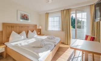 A bright bedroom with a double bed and two large pillows. The room has windows with curtains and a small table.