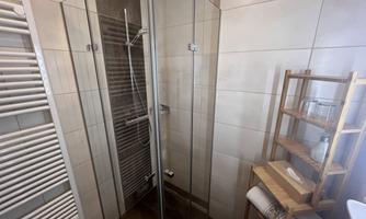 A modern shower with glass walls and a minimalist wooden shelf in the bathroom. The walls are light-tiled and create a clean atmosphere.
