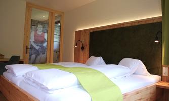 A cozy bedroom with a large bed, white pillows, and a green blanket. In the background, there is a glass door through which a piece of artwork is visible.