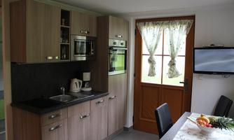 A modern kitchen with wooden cabinets and a small dining table. The room has large windows with curtains and a television on the wall.