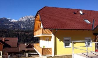 A modern house with a red roof and balconies. In the background, majestic mountains and a blue sky can be seen.