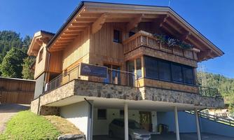 A modern wooden house with a large balcony and stone base. It is situated in a picturesque mountain landscape, surrounded by trees.