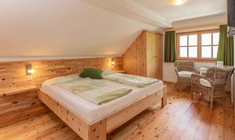 A cozy bedroom with a large double bed and wood paneling. There is a seating area with two chairs and a table, as well as windows with green curtains.