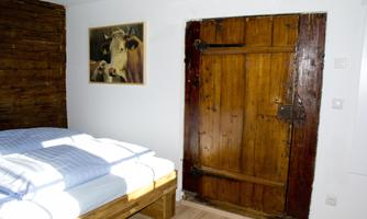 A cozy bedroom with a wooden bed and fresh white bed linens. A picture of a cow hangs on the wall, and an old wooden door is visible.