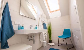 A modern bathroom with bright tiles and a large window. Decorative glasses are placed on the sink, and a blue chair complements the decor.