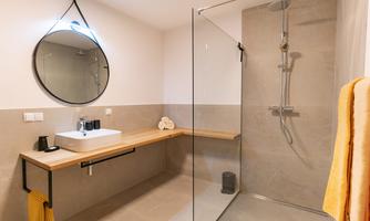 A modern bathroom with a glass shower and an elegant vanity. A round mirror hangs on the wall and a towel in yellow is ready.