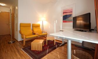 A modern living room with two yellow armchairs, a table, and a television. The room has wooden flooring and a cozy atmosphere.
