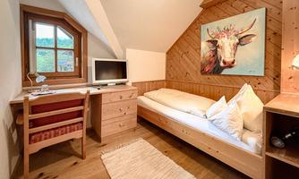 A cozy room with wooden paneling and a single bed. A piece of art featuring a cow hangs on the wall, and a desk stands next to a window.