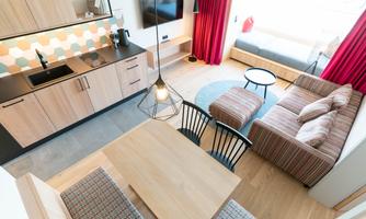 A modern kitchen and living area combination with wooden surfaces and red curtains. There is a dining area with a table and comfortable chairs, as well as a sofa for relaxing.