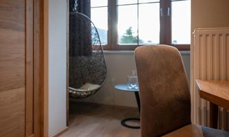 A cozy room with a chair, a table, and a modern hanging chair next to the window. The wooden floor and the warm colors create an inviting atmosphere.