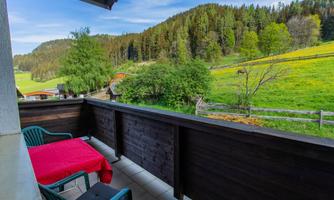 A beautiful balcony with a red table and two chairs. In the background, green meadows and forests can be seen.