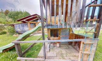 A playground with a wooden structure, a slide, and a ladder. The area is surrounded by grass and has a rustic atmosphere.