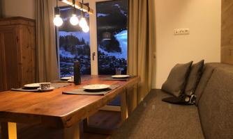 A cozy dining area with a large wooden table and comfortable seat cushions. The windows offer a picturesque view of the mountains.