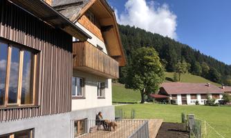 A modern house with wooden elements and a balcony, surrounded by green meadows and wooded hills. In the background, additional buildings and blue sky can be seen.