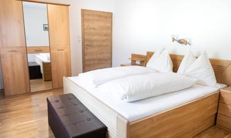 A bright bedroom with a large double bed and two pillows. In the background, there is a mirror and a wooden wardrobe.