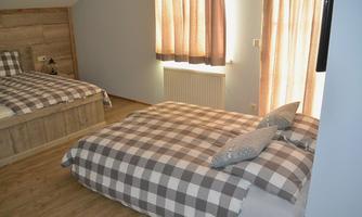 A cozy bedroom with two beds, equipped with checkered bedspreads and pillows. The room has light-colored curtains and wooden floors.
