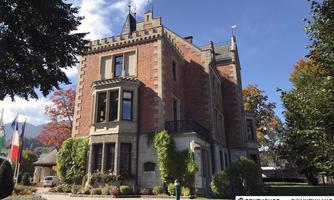An impressive castle with a red brick design and distinctive towers. The garden is well-maintained and surrounded by colorful foliage.