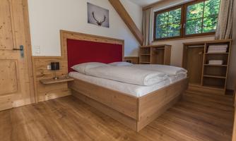 A cozy bedroom with a wooden bed and red accents. Large windows allow for plenty of natural light and offer a view of the greenery.