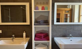 A modern bathroom with two sinks and large mirrors. In the center, colored towels and utensils are neatly arranged on a shelf.