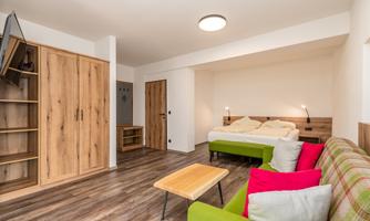 A modern room with a double bed and a cozy seating area. The room features wooden shelves and a television on the wall.