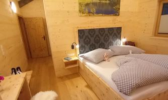 A cozy bedroom with wooden walls and a pine bed and a large window. The bed is equipped with checkered pillows and a blanket, and a beautiful picture hangs above the bed.