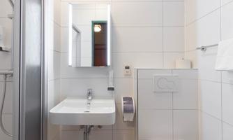 A modern bathroom with a sink, a mirror, and a shower. The walls are light tiled and the lighting is pleasant.