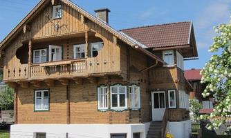 A traditional wooden house with a charming balcony and many windows. It is situated in a green environment with flowers and a clear sky.