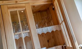 A wooden kitchen cabinet with glasses and a white lace border. Below is a teapot and cups.