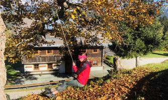 A girl swings on a rope beneath a tree, surrounded by colorful autumn leaves. In the background, a traditional wooden house can be seen.